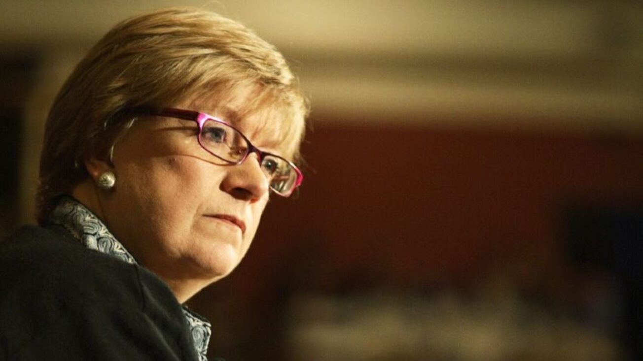 A woman with short blonde hair and glasses looks aslant across the image