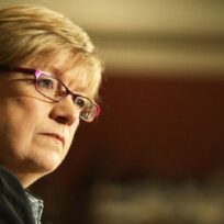 A woman with short blonde hair and glasses looks aslant across the image