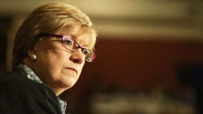 A woman with short blonde hair and glasses looks aslant across the image