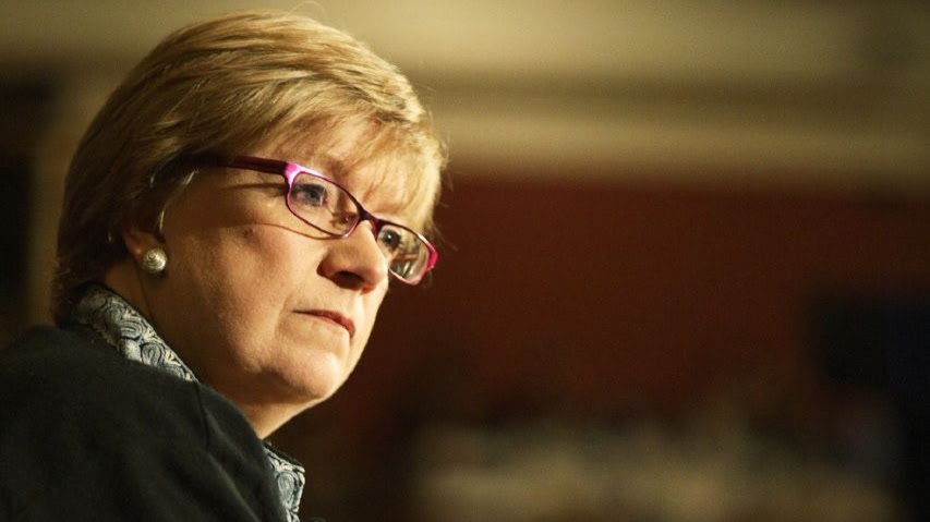 A woman with short blonde hair and glasses looks aslant across the image