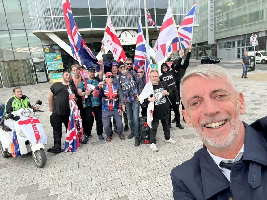 A group of people carrying St George's Cross and Union Jack flags behind a man with grey hair and beard taking a selfie