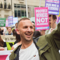 A man in a trenchcoat waves with placards in the background