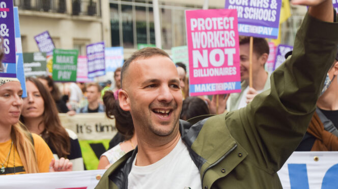 A man in a trenchcoat waves with placards in the background