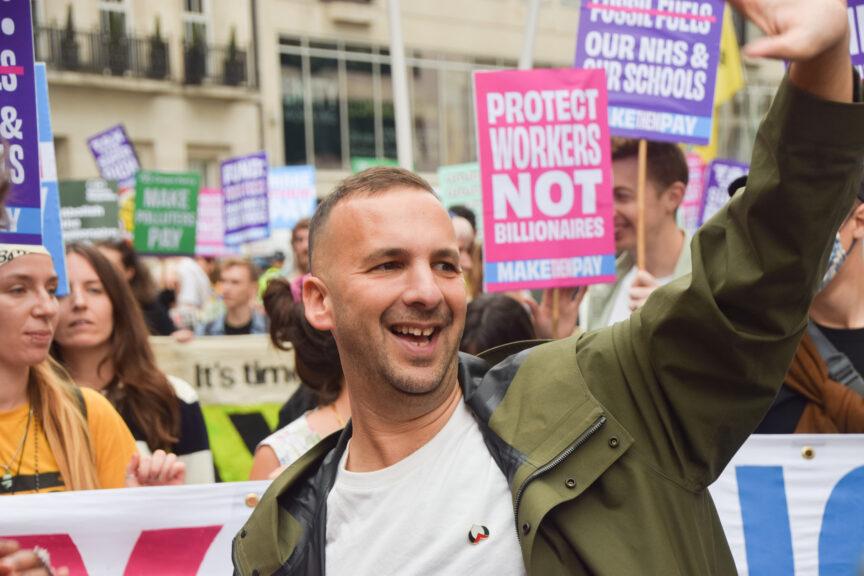 A man in a trenchcoat waves with placards in the background