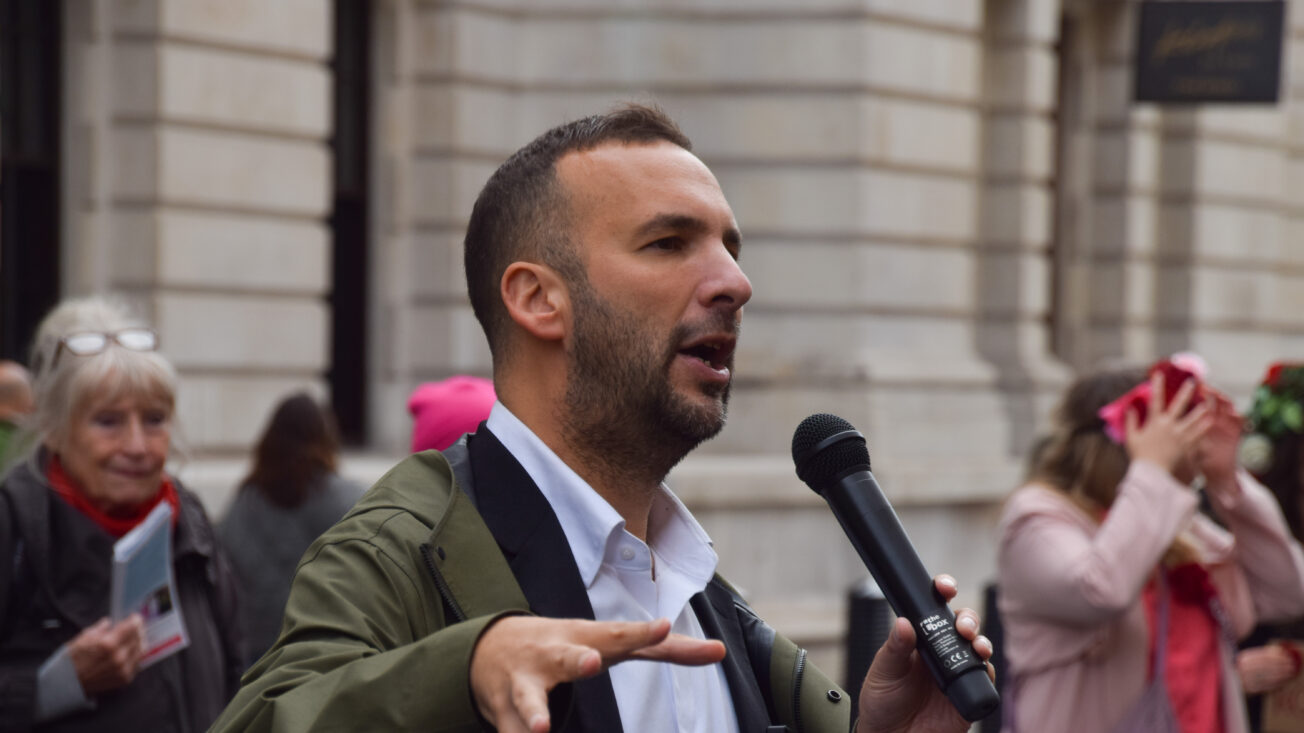 A man wearing a white shirt and a green rainjacket speaks into a mic