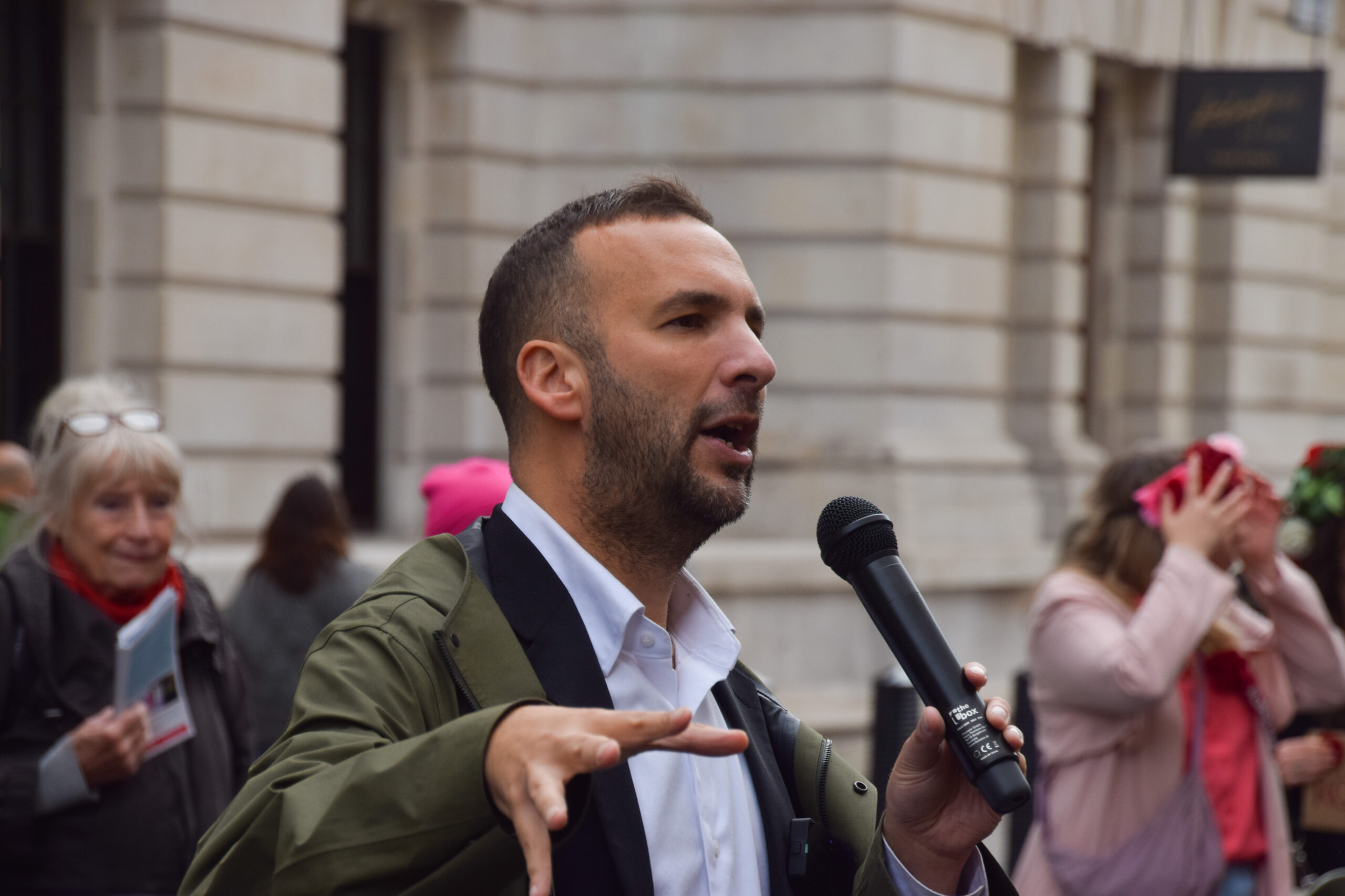 A man wearing a white shirt and a green rainjacket speaks into a mic
