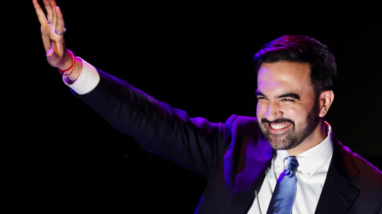 A man in a suit smiles and waves against a black background