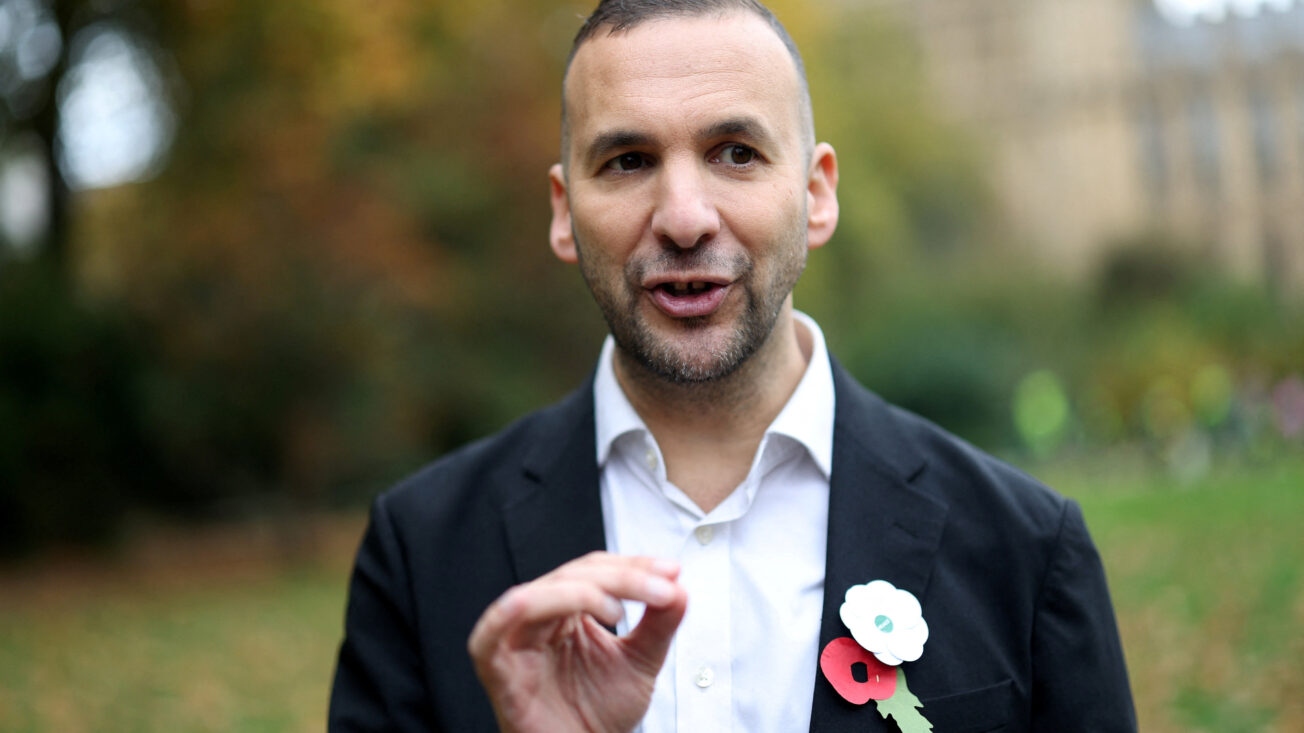 A man with short brown hair in a suit wears a red and white poppy together