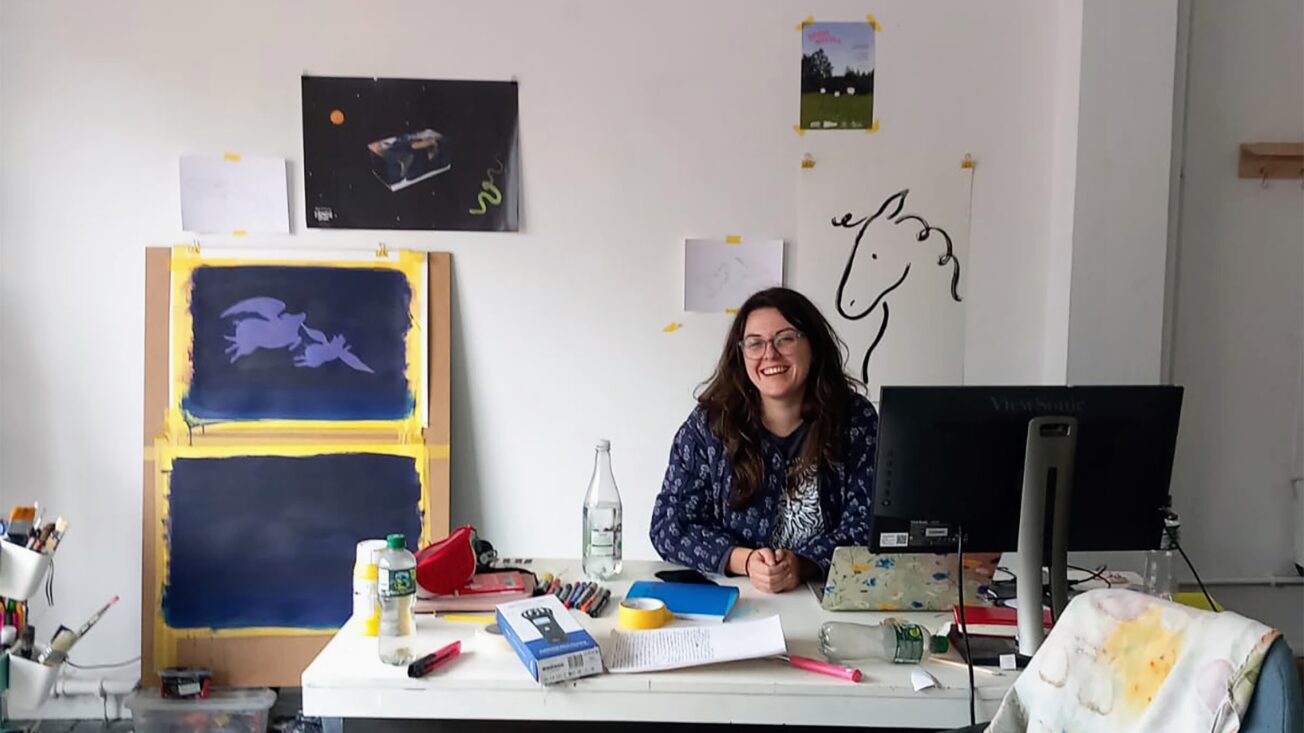 A woman with long black hair and black glasses sitting at a desk amid pieces of artwork.
