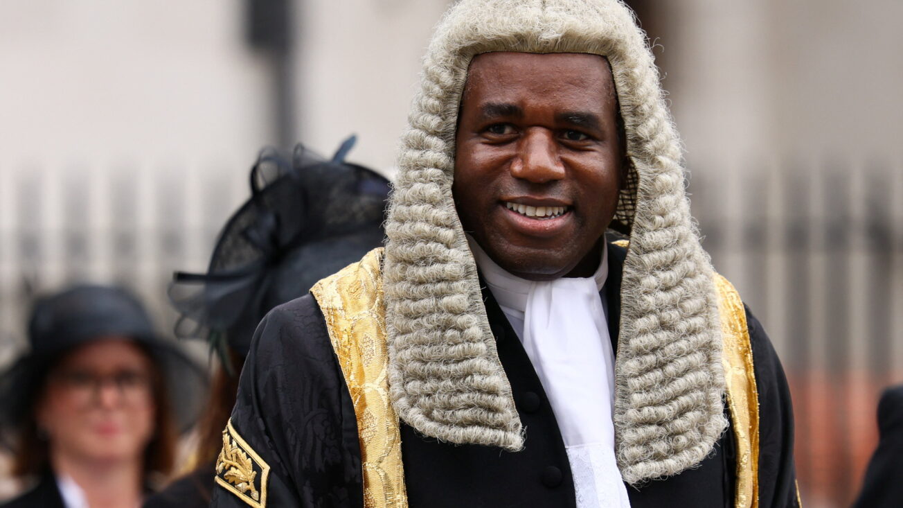A man in a wig and black and gold velvet coat smiles