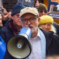 A man with a grey beard, beige baseball cap and glasses holds a loudhailer as people holding placards stand behind him