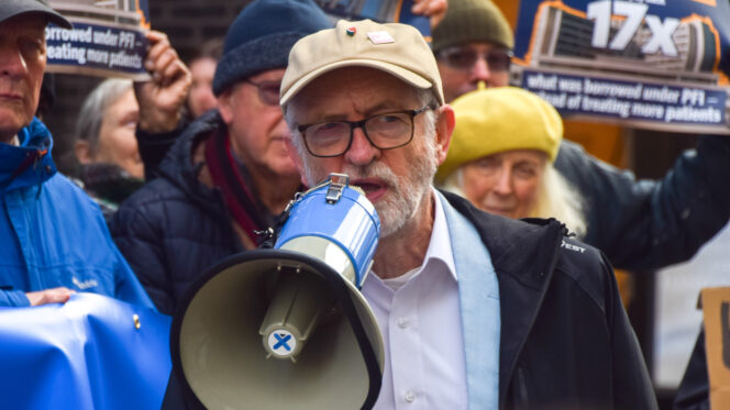A man with a grey beard, beige baseball cap and glasses holds a loudhailer as people holding placards stand behind him