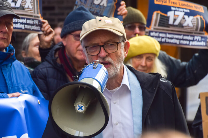 A man with a grey beard, beige baseball cap and glasses holds a loudhailer as people holding placards stand behind him