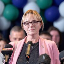 A woman with blonde hair, black glasses and a grey top stands in front of several microphones, as people and balloons are behind her