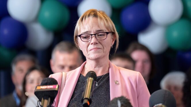 A woman with blonde hair, black glasses and a grey top stands in front of several microphones, as people and balloons are behind her