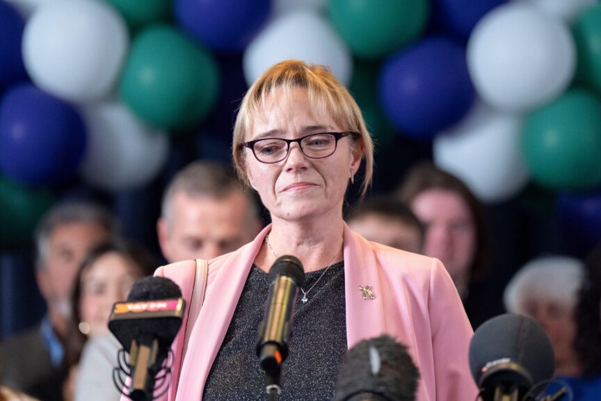 A woman with blonde hair, black glasses and a grey top stands in front of several microphones, as people and balloons are behind her