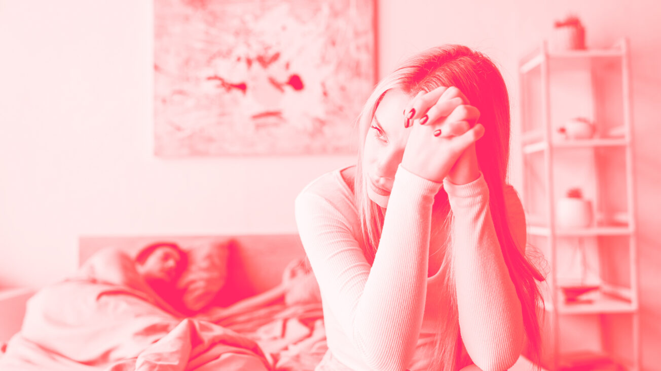 A woman looks into the distance as a man lies in bed topless