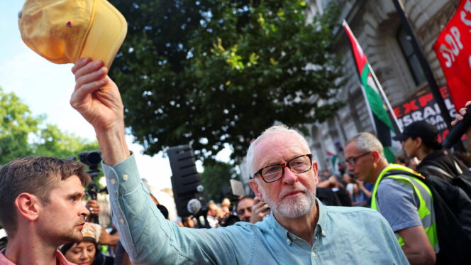 A man with grey hair and glasses, wearing a blue shirt, waves a yellow baseball cap above his head