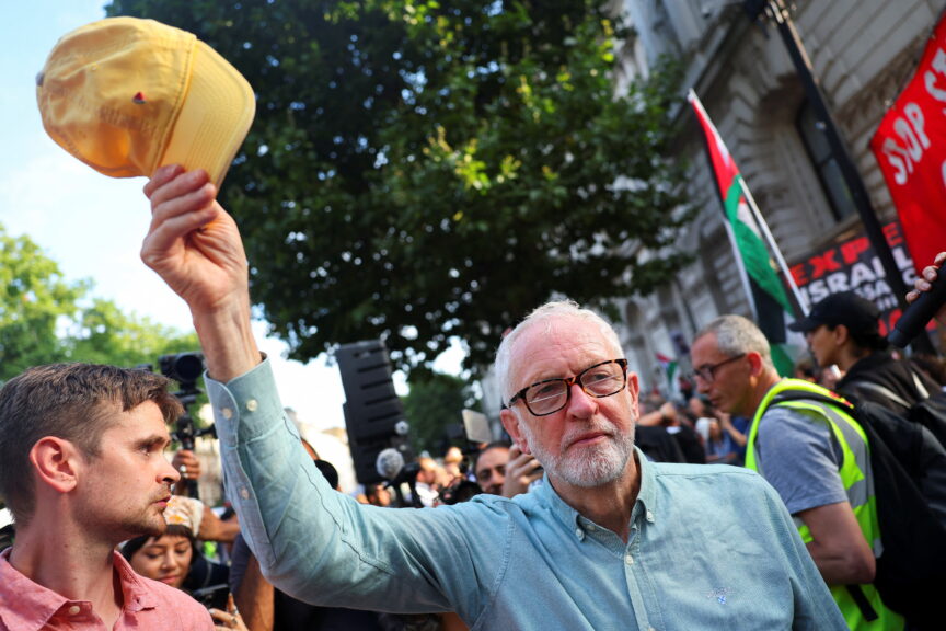 A man with grey hair and glasses, wearing a blue shirt, waves a yellow baseball cap above his head