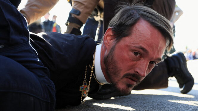 A red-faced man wearing a crucifix is held to the ground by a police officer