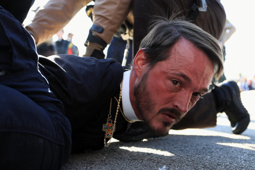 A red-faced man wearing a crucifix is held to the ground by a police officer