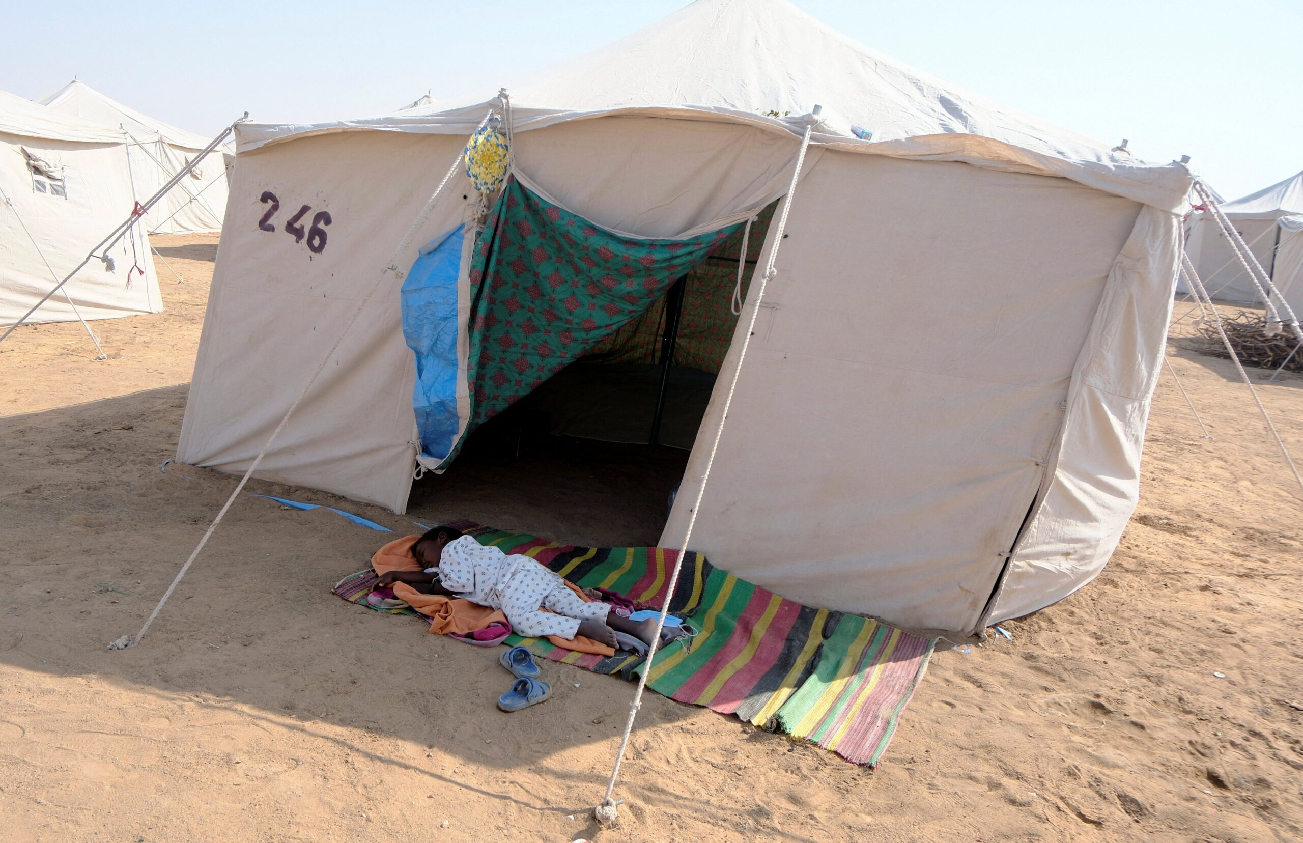 A child from El Fasher sleeps on the dusty ground outside a tent.