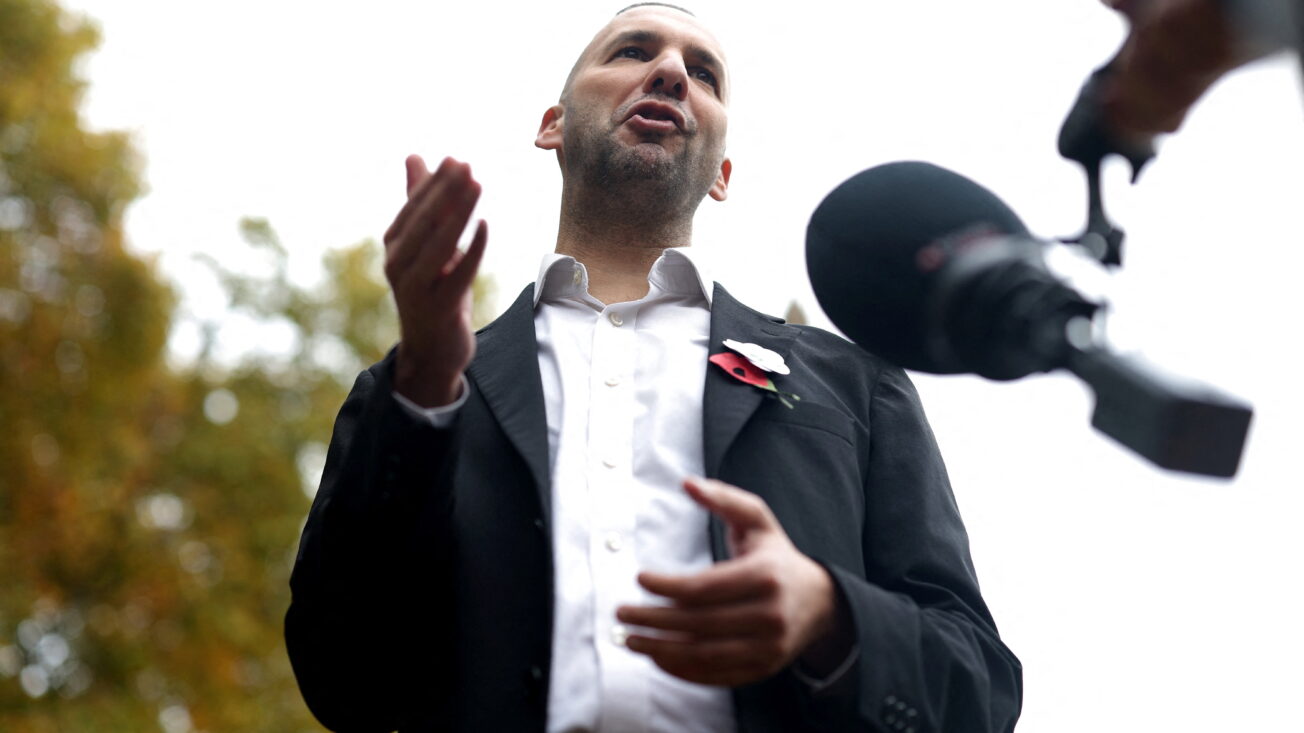 A man in a black suit and white shirt stands in front of a microphone, shot from below