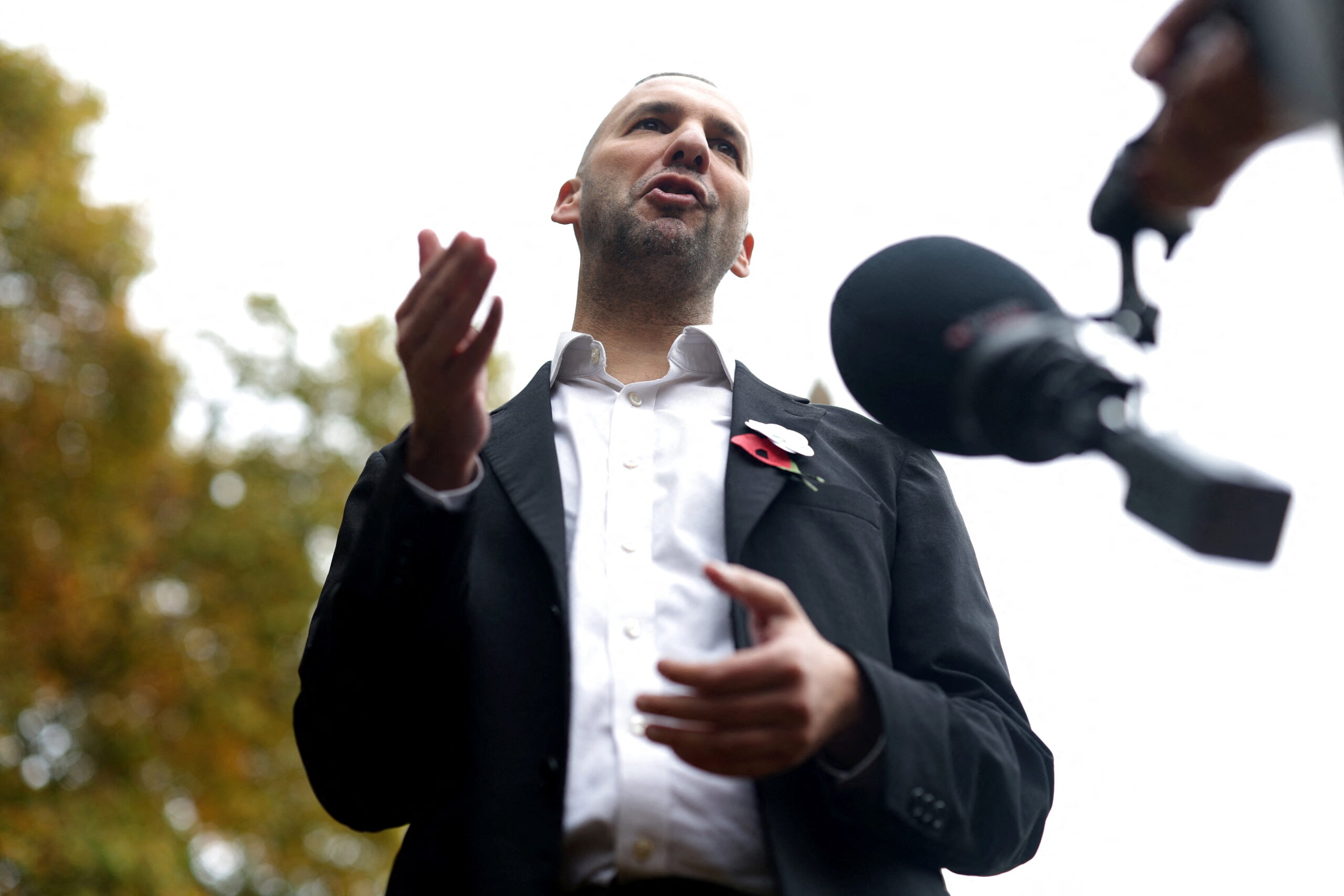 A man in a black suit and white shirt stands in front of a microphone, shot from below