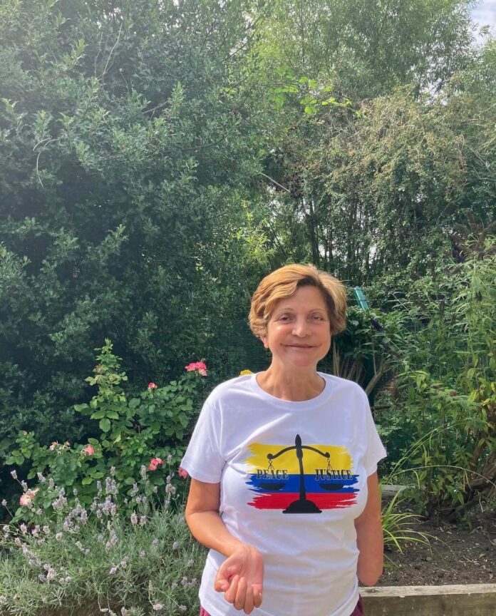 A woman with brown hair, a white t-shirt with 'peace and justice' written on it poses in front of a garden scene