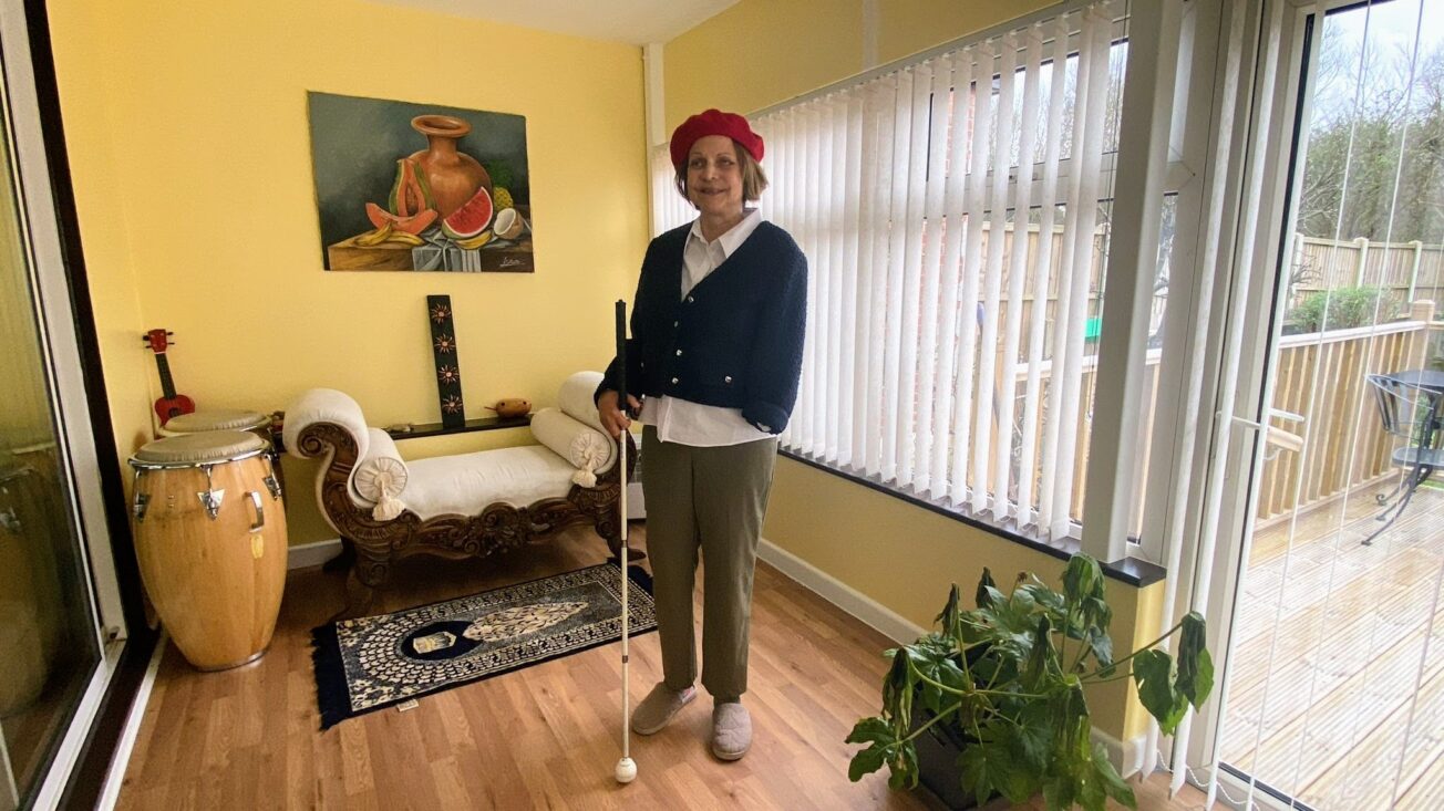 A woman with shoulder-length brown hair and a red beret holds a cane in her right hand. Her left arm is amputated. She stands in a yellow living room with a chaise longue and a still life painting. A drum stands in the corner. Through the blinds we can see a wooden patio